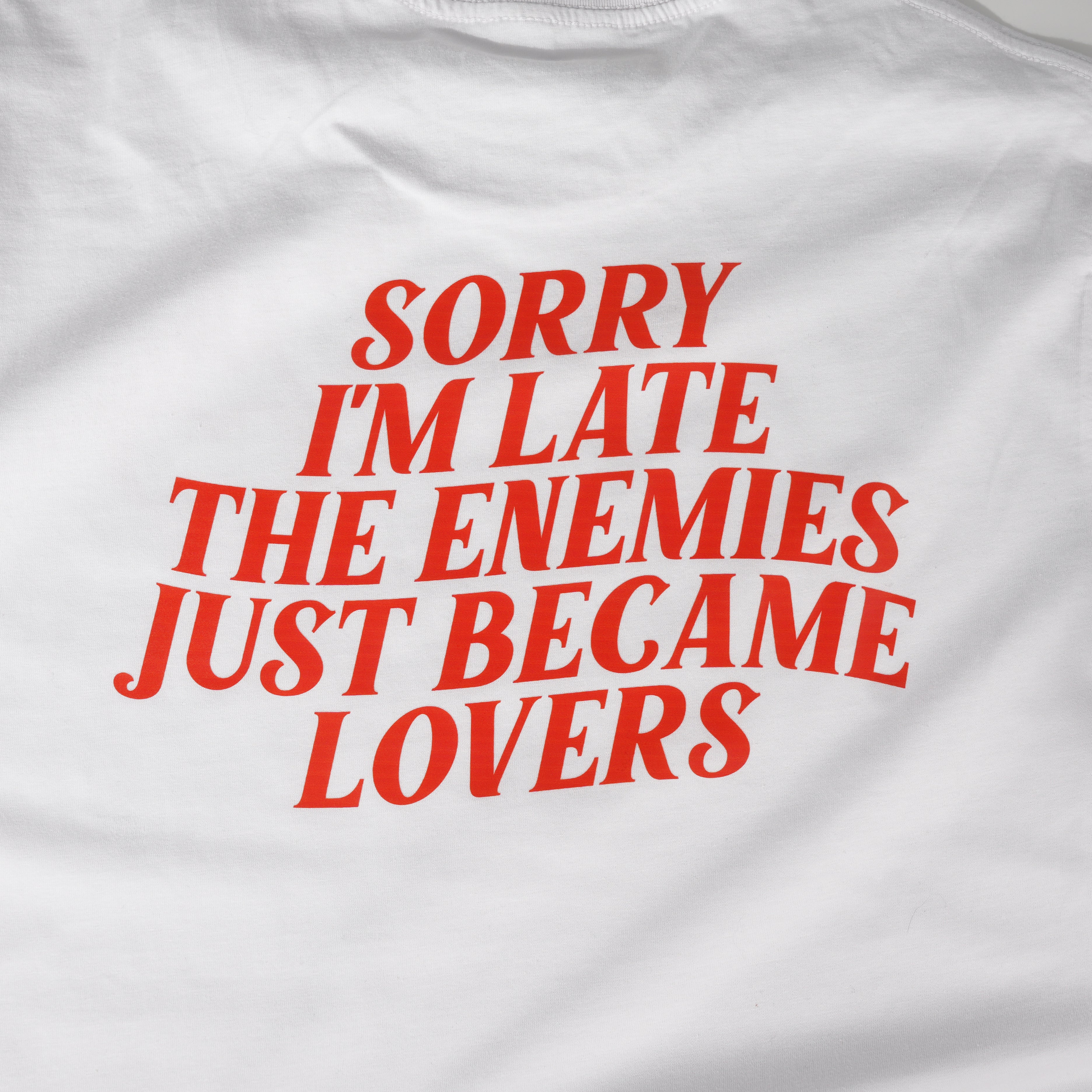 White t-shirt with red text 'Sorry I'm late. The enemies just became lovers' on a white background